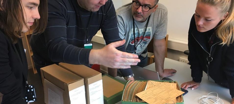 Tre menn og en kvinne står rundt et et bord mens de ser i en eske med papirer.