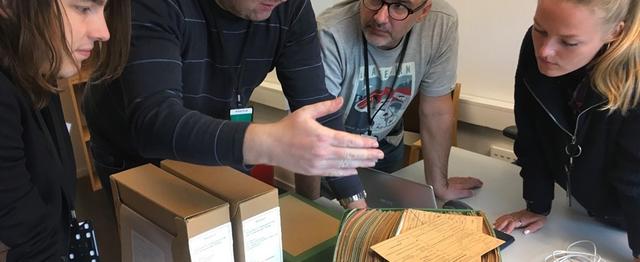 Tre menn og en kvinne står rundt et et bord mens de ser i en eske med papirer.