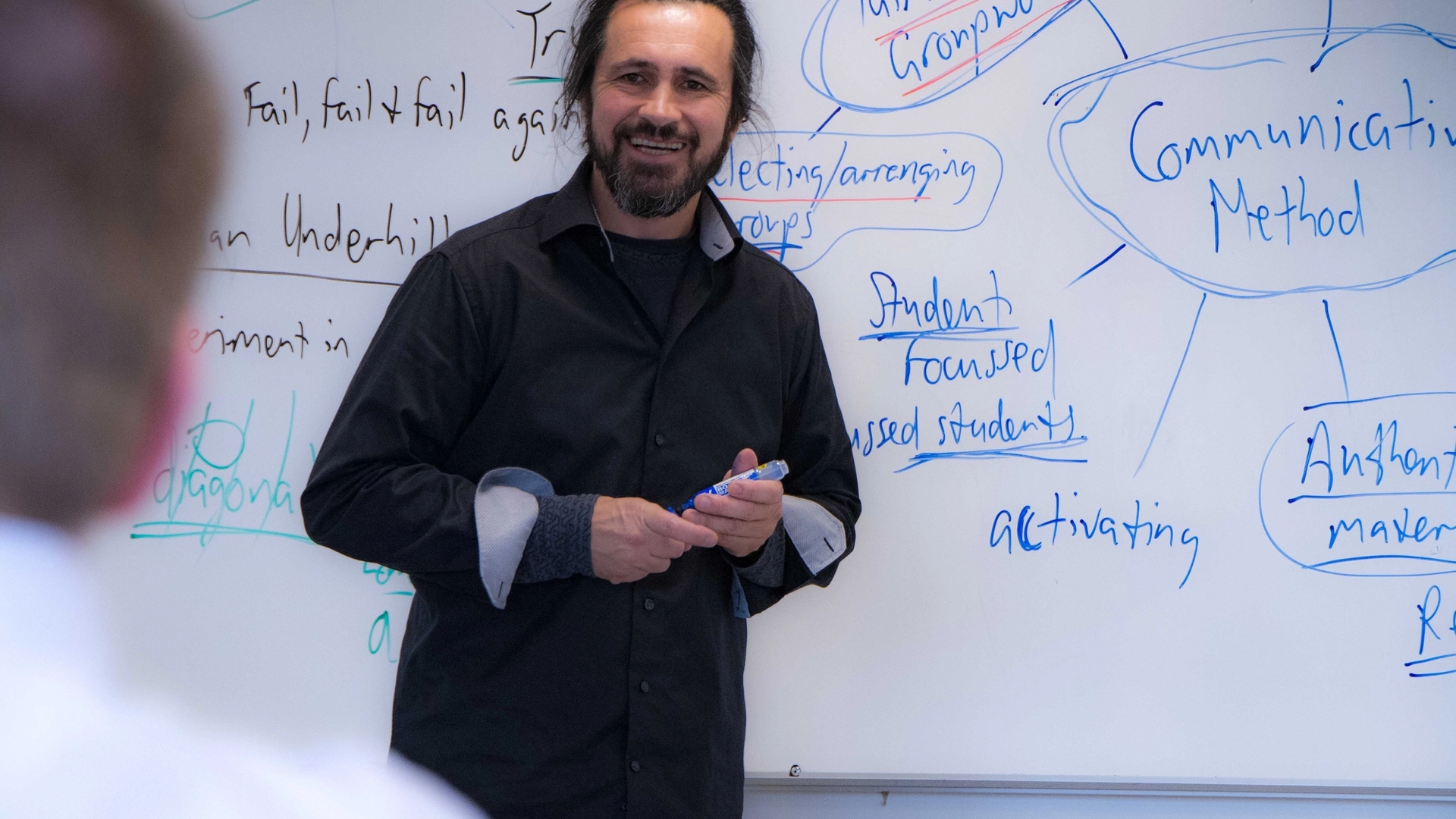 Mann står foran en tavle mens han holder en tusj og smiler mot en klasse.