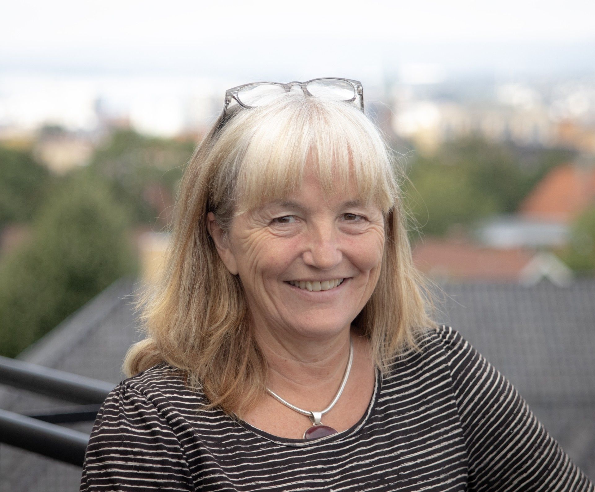 Torunn Thomassen smiler mot kameraet. Hun er fotografert utendørs med uskarp bybebyggelse i bakgrunnen.