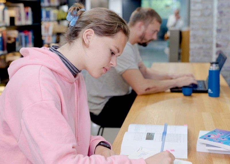 To studenter på campus. Kvinne som ser i lærebøker, mann i bakgrunnen ser på laptop