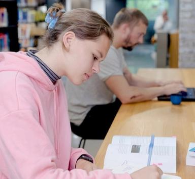 To studenter på campus. Kvinne som ser i lærebøker, mann i bakgrunnen ser på laptop