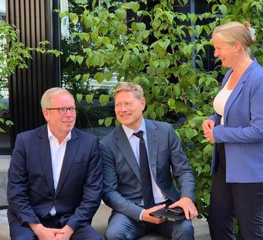 To menn i dress sitter ute på en mur, mens en kvinne i blå jakke står ved siden av.