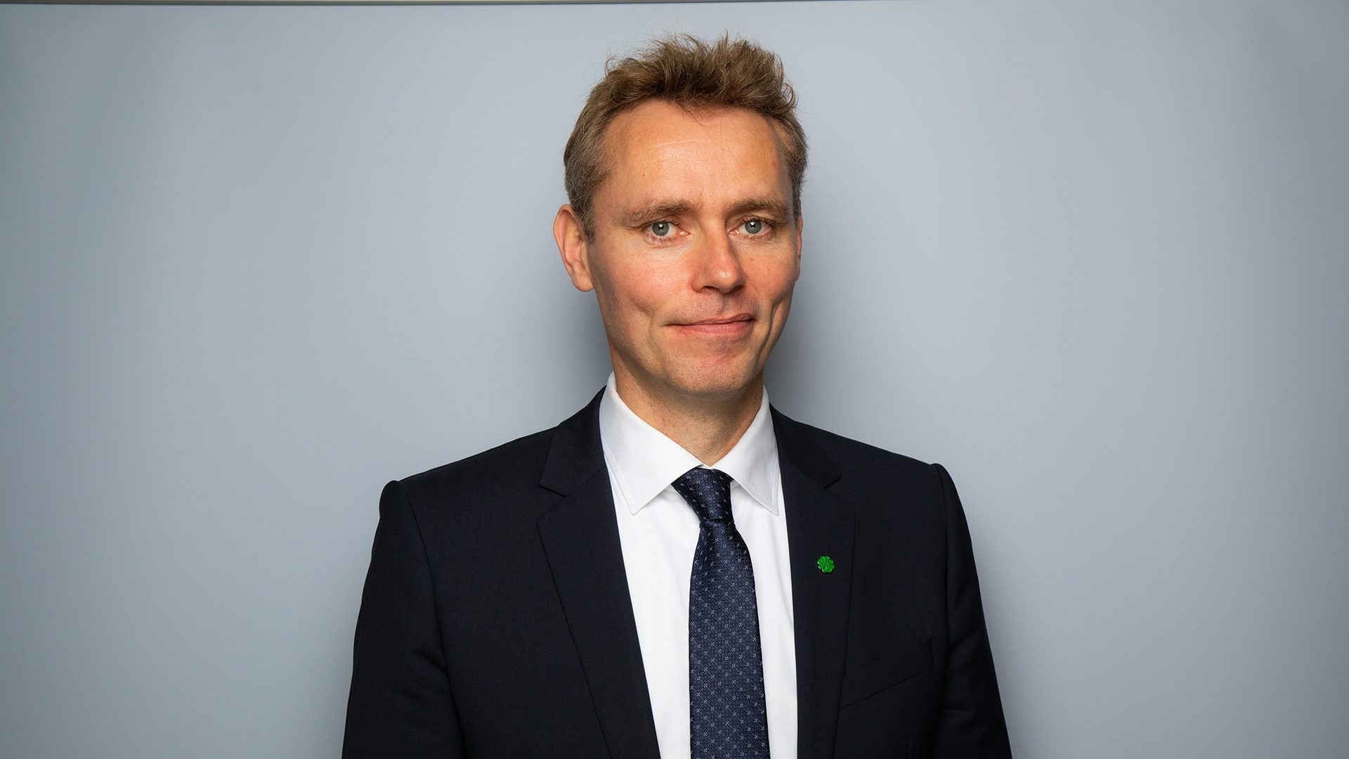 Mann i hvit skjorte, blå-grått slips og svart dressjakke ser i kamera