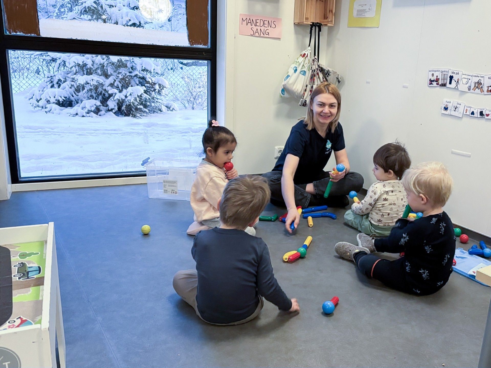 Barnehagelærer sitter på gulvet sammen med flere små barn i barnehage og spiller med fargerike mikrofoner. Barna leker og utforsker lyder innendørs, med snødekt landskap synlig gjennom vinduet i bakgrunnen.