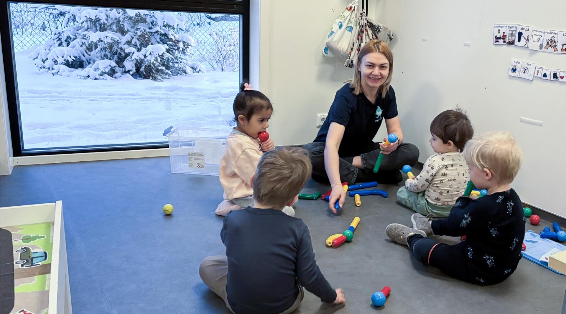 Barnehagelærer sitter på gulvet sammen med flere små barn i barnehage og spiller med fargerike mikrofoner. Barna leker og utforsker lyder innendørs, med snødekt landskap synlig gjennom vinduet i bakgrunnen.