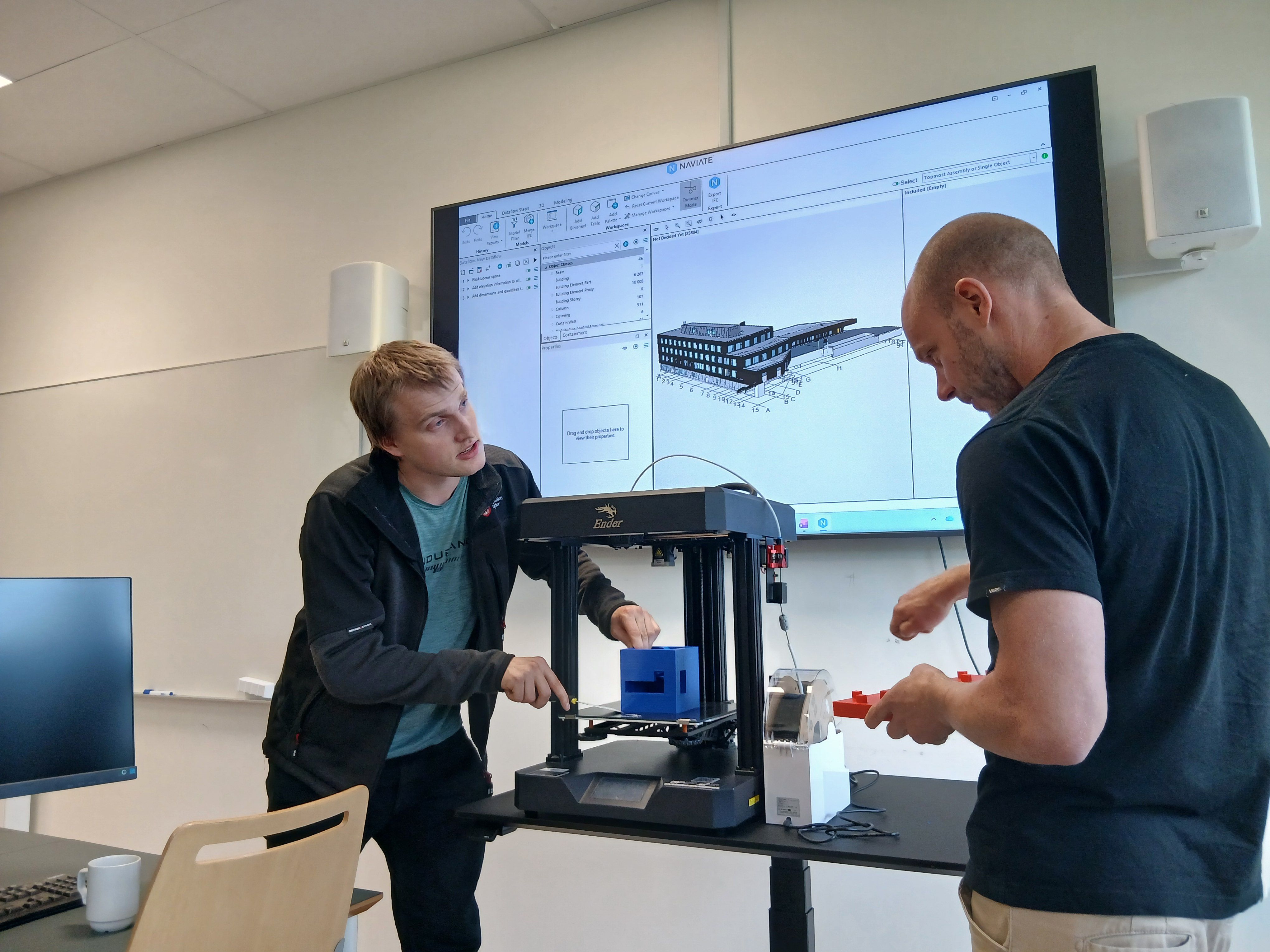 Kristian Spikkeland og Audun Slåtterø demonstrerer 3D-printing i eit laboratorium ved Fagskulen i Agder. Ein digital BIM-modell av eit bygg er synleg på skjermen i bakgrunnen.