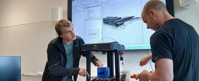 Kristian Spikkeland og Audun Slåtterø demonstrerer 3D-printing i eit laboratorium ved Fagskulen i Agder. Ein digital BIM-modell av eit bygg er synleg på skjermen i bakgrunnen.