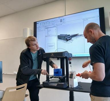 Kristian Spikkeland og Audun Slåtterø demonstrerer 3D-printing i eit laboratorium ved Fagskulen i Agder. Ein digital BIM-modell av eit bygg er synleg på skjermen i bakgrunnen.