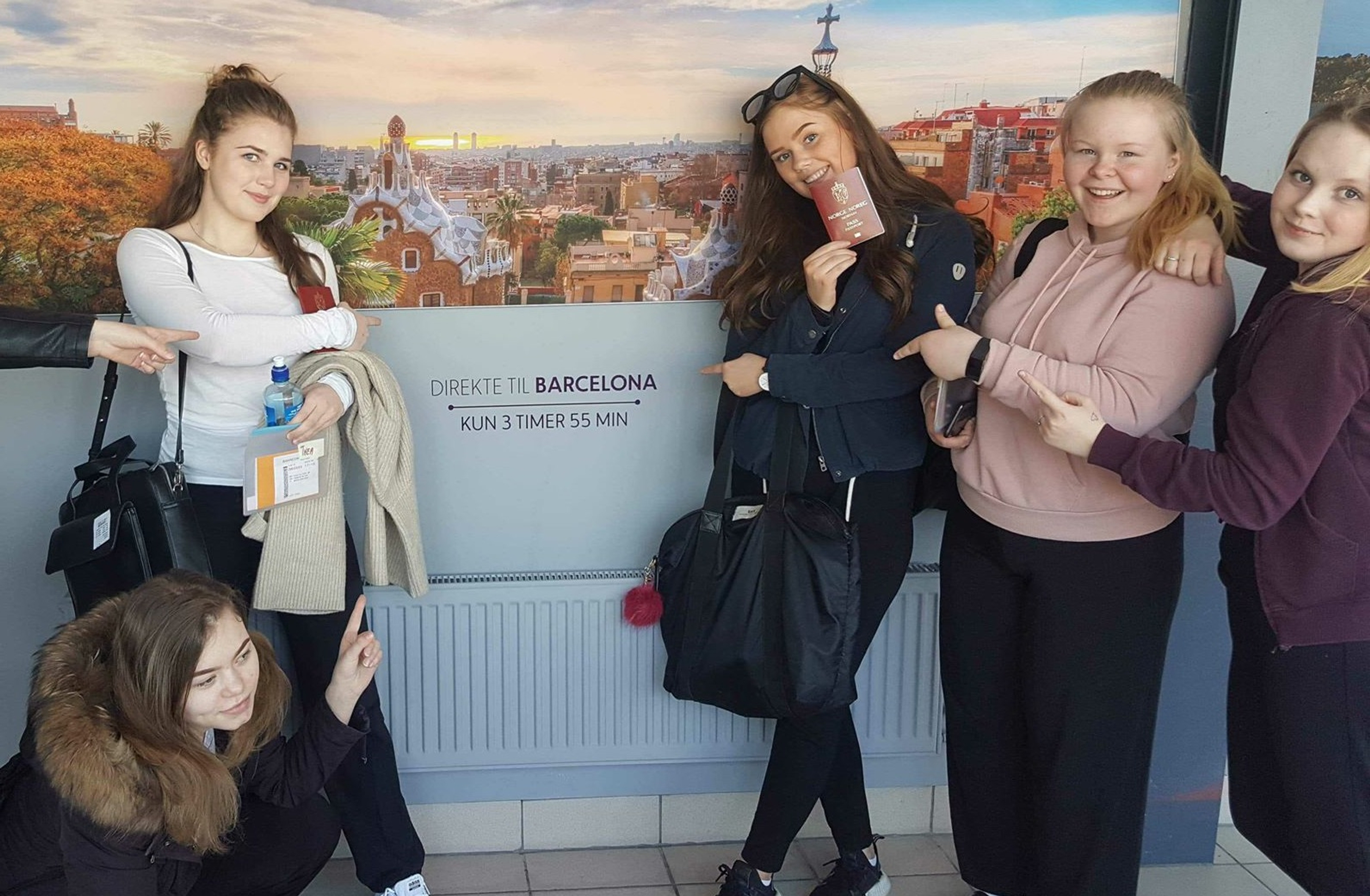 Fem unge jenter står foran et skilt på en flyplass og peker på skiltet som sier at det er 3 timer til Barcelona.