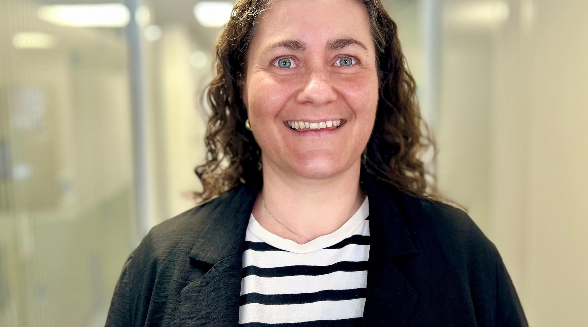 Kvinne med krøllete mørkt hår. Hvit og svart stripede t-skjorte og en svart cardigan. Ser i kamera og smiler.