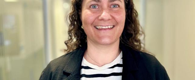 Therese Røst, kvinne med krøllete mørkt hår. Hvit og svart stripede t-skjorte og en svart cardigan. Ser i kamera og smiler.