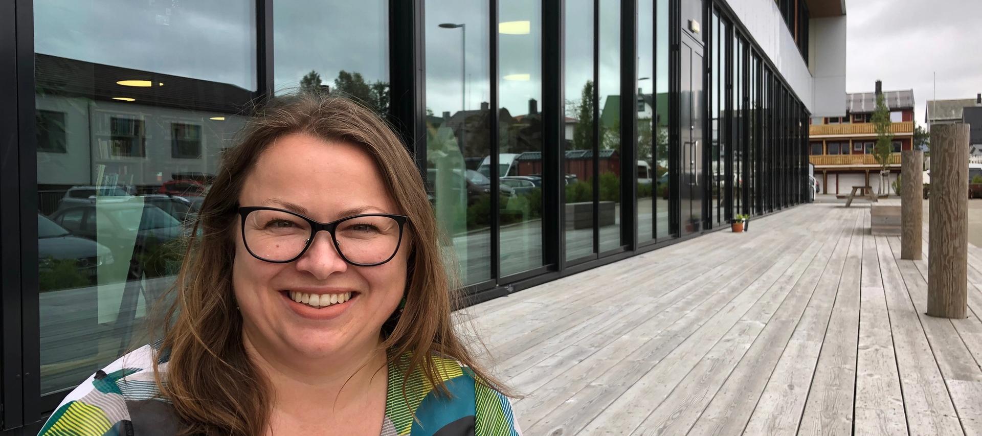 Kvinne med briller smiler mot kamera. Er utendørs foran en bygning med glassfasade.
