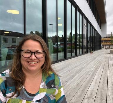 Kvinne med briller smiler mot kamera. Er utendørs foran en bygning med glassfasade.