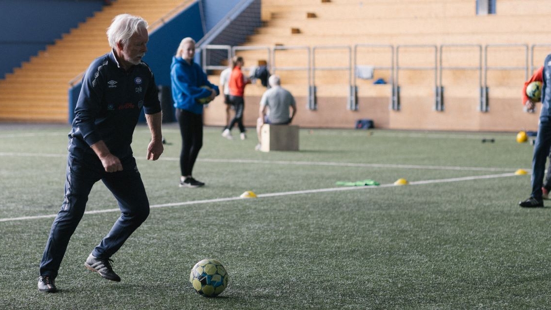 Flere fotballspillere på en bane. Eldre mann i fokus mens han skal til å sparke en ball.