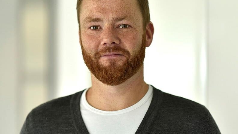 Mann med skjegg, grå genser og hvit t-skjorte som ser i kamera med et litt skeptisk blikk.
