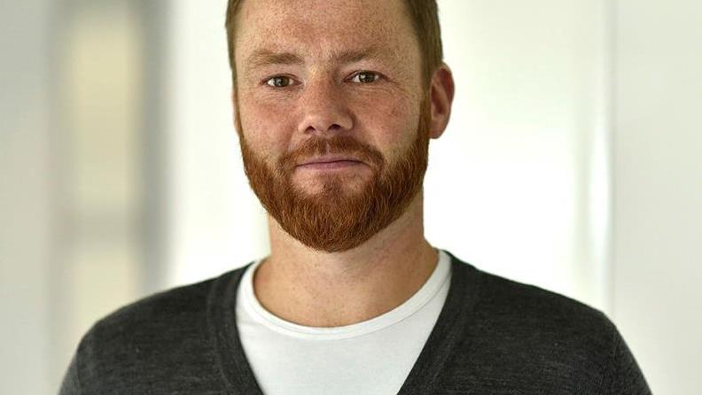 Mann med skjegg, grå genser og hvit t-skjorte som ser i kamera med et litt skeptisk blikk.