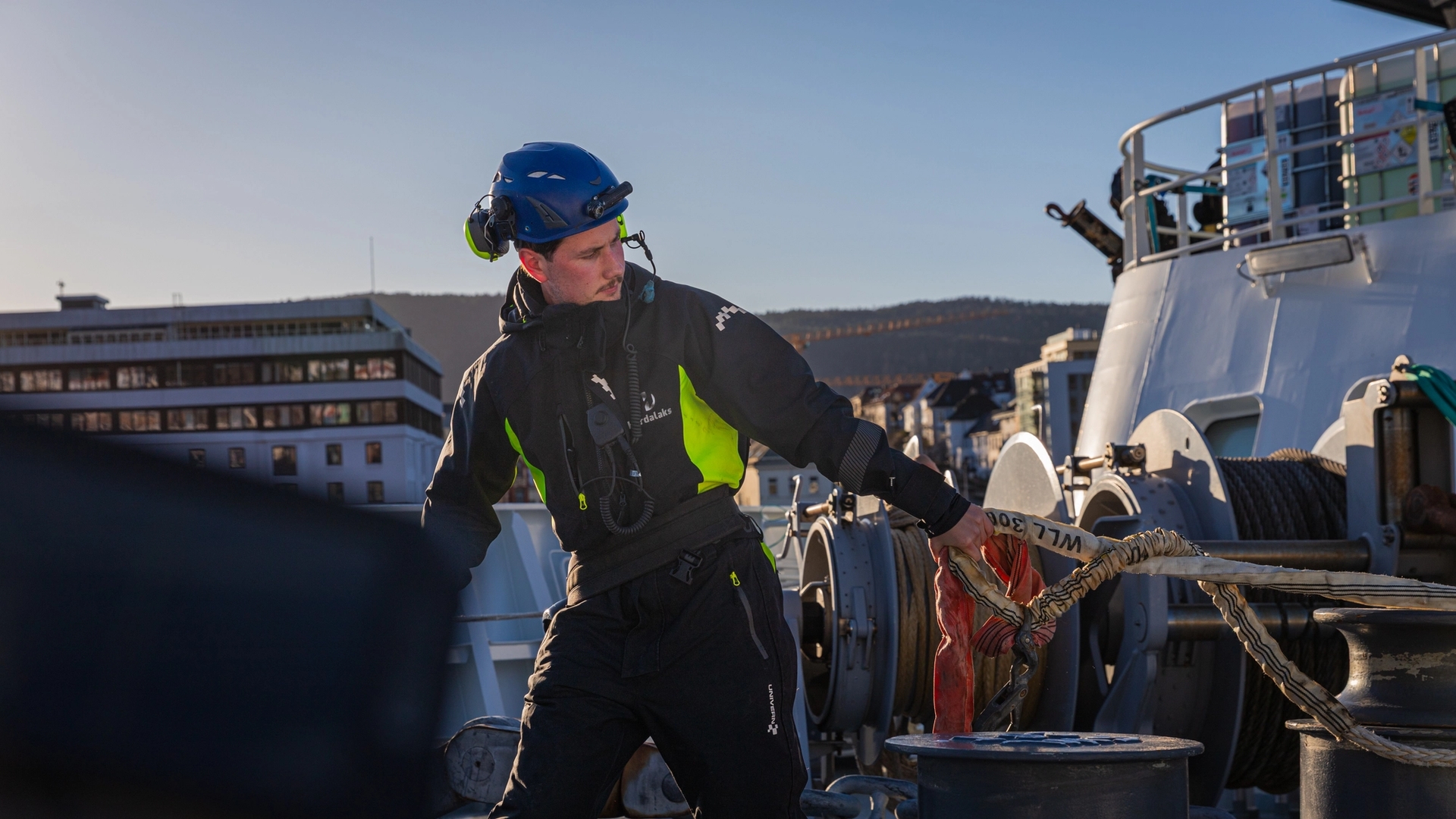 Mann i verneutstyr arbeider med tau og utstyr på dekk av et skip i havn.