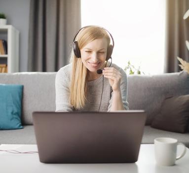 Smilende kvinne sitter i sofa med en PC foran seg og har på seg et hodesett med mikrofon.