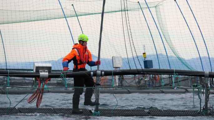 Mann med hjelm og redningsvest balanserer på kanten av merden ute på havet