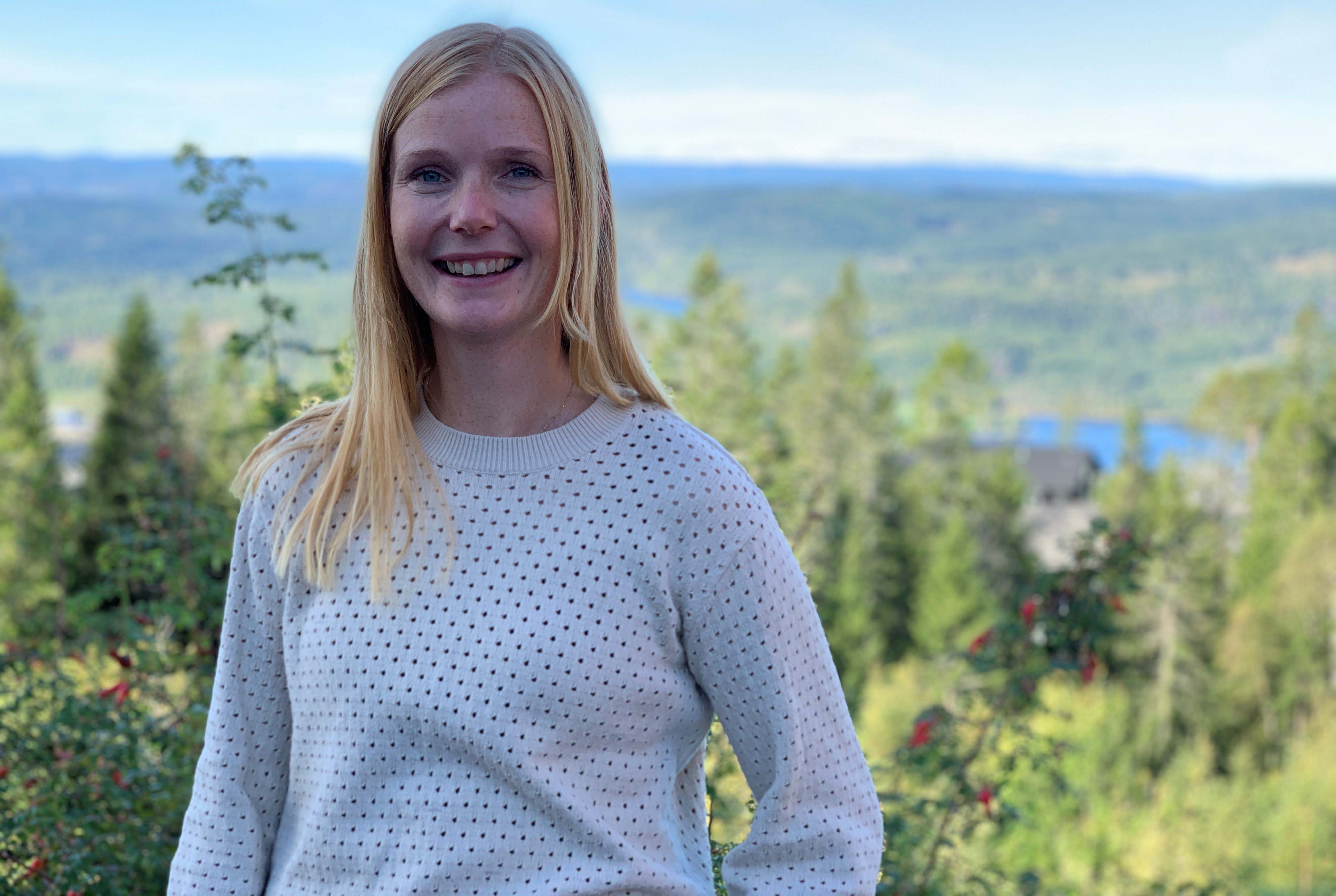 Kvinnelig lærer med langt lyst hår og hvit genser. Står ute i naturen og smiler mot kamera.