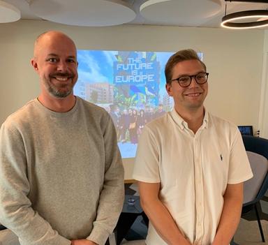 To menn står foran et lerret mens de smiler mot kamera.