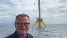 Mann med briller smiler foran en vindmølle på havet