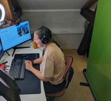Kvinnelig lærer fjernunderviser fra studio med tre skjermer, ringlys og grønnskjerm.