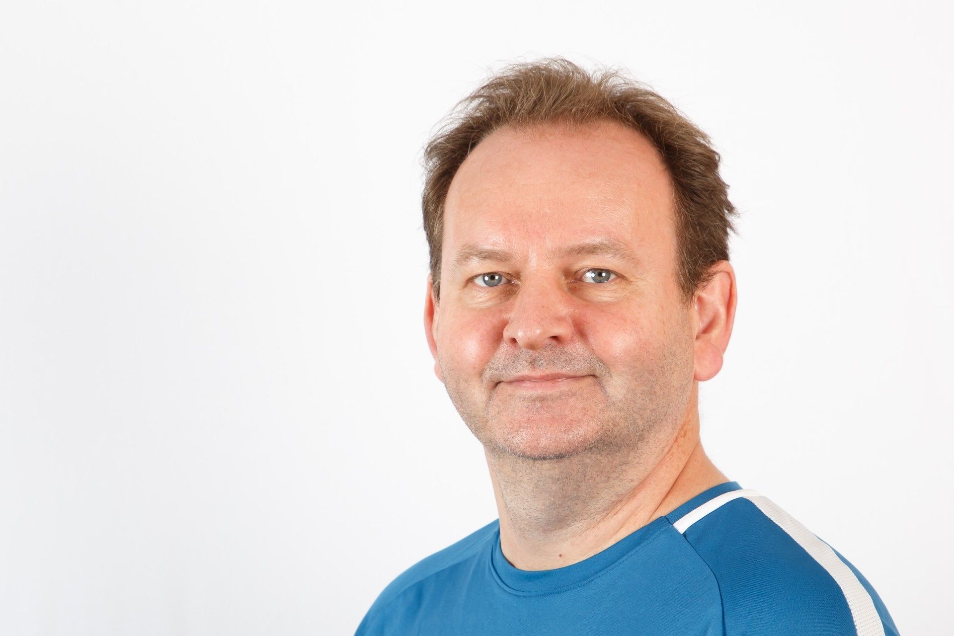 Portrett av en mann med kort brunt hår og blå T-skjorte, mot en lys, nøytral bakgrunn.