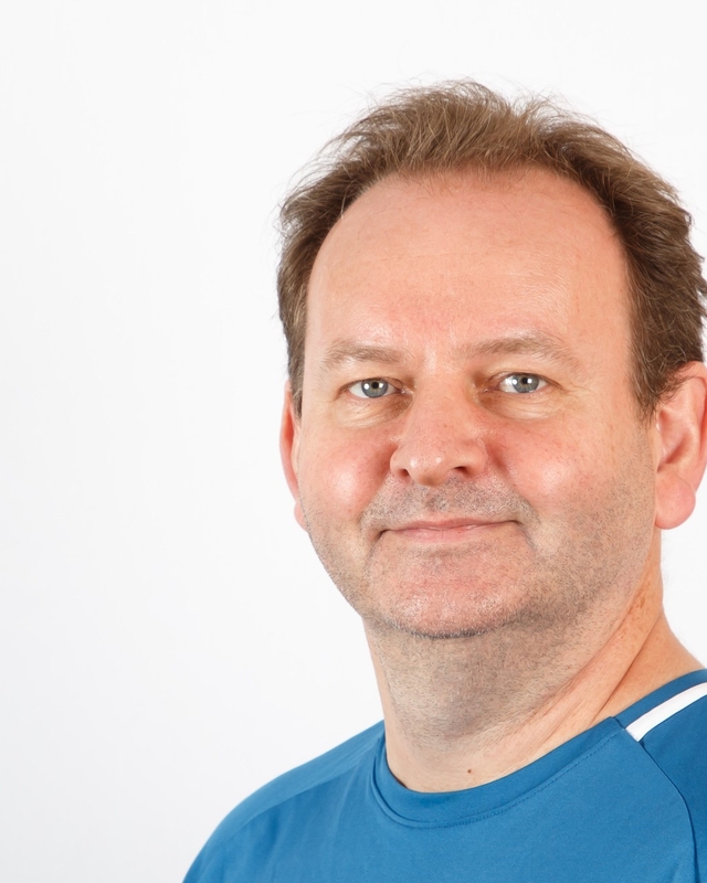 Portrett av en mann med kort brunt hår og blå T-skjorte, mot en lys, nøytral bakgrunn.