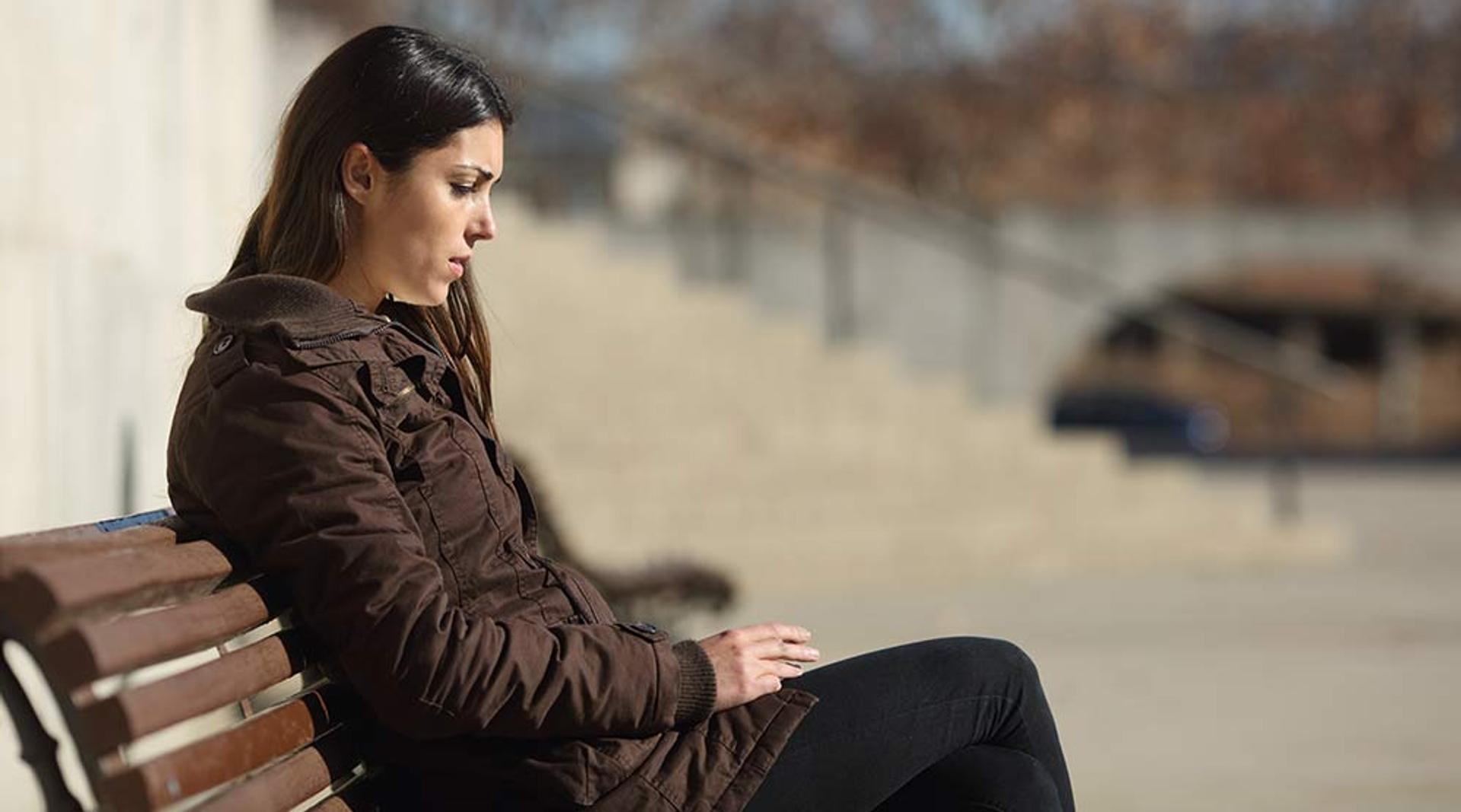 Jente med mørkt hår som sitter på en benk. Har på seg brun jakke og sort bukse. Ser trist ut. 