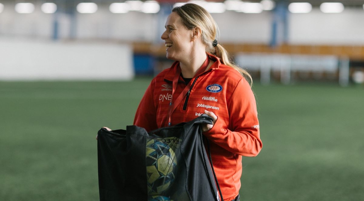 Kvinne i treningsklær smiler mens hun holder et nett med fotballer.