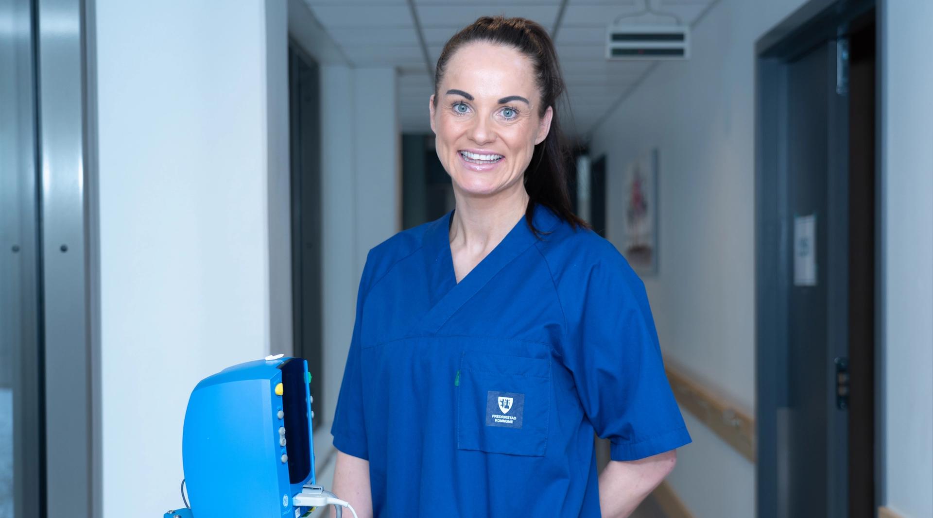 Person i blå arbeidsuniform står i en korridor i en helseinstitusjon ved siden av medisinsk utstyr.
