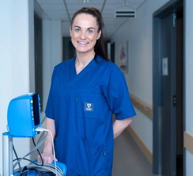 Person i blå arbeidsuniform står i en korridor i en helseinstitusjon ved siden av medisinsk utstyr.