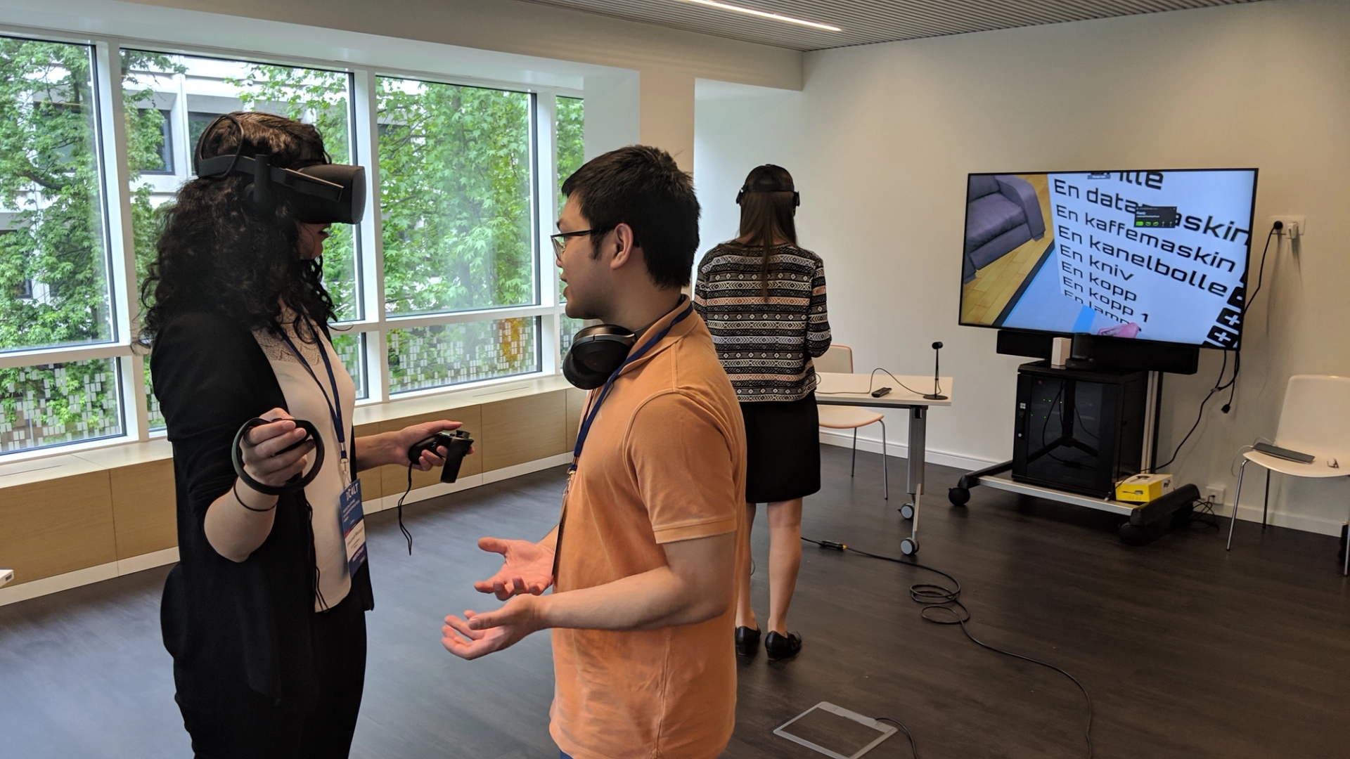Kvinner prøver VR-briller mens en mann forklarer mens en mann snakker med henne. Bak ser man en annen kvinne med VR-briller og en TV-skjerm.