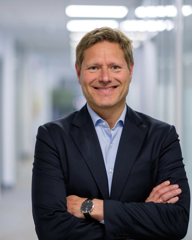 Mann med sort dress og lyseblå skjorte som ser i kamera med armene i kors, mens han smiler