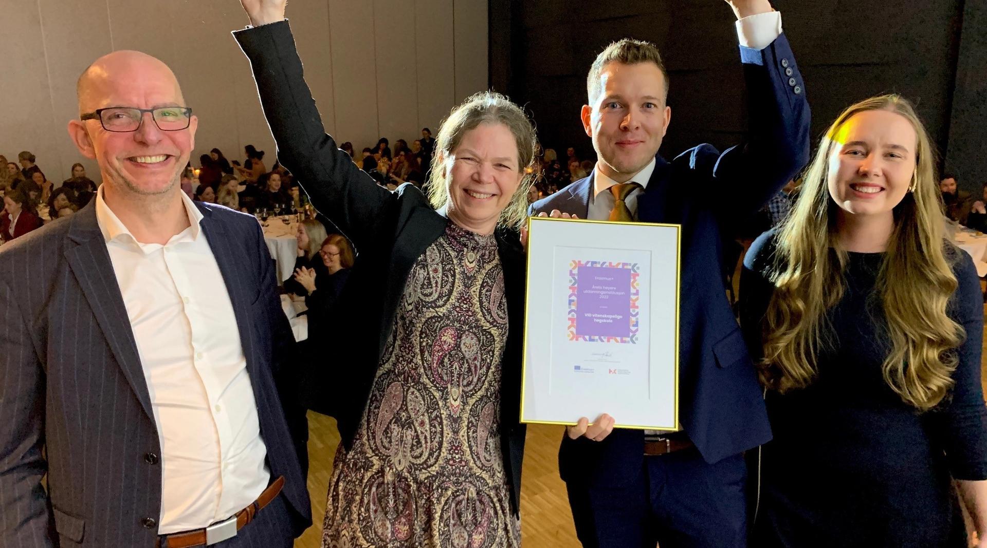 To kvinner og to menn smiler glad mot kamera. Kvinne og mann i midten holder hendene i luften mens de stolt viser frem et diplom.