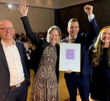 To kvinner og to menn smiler glad mot kamera. Kvinne og mann i midten holder hendene i luften mens de stolt viser frem et diplom.