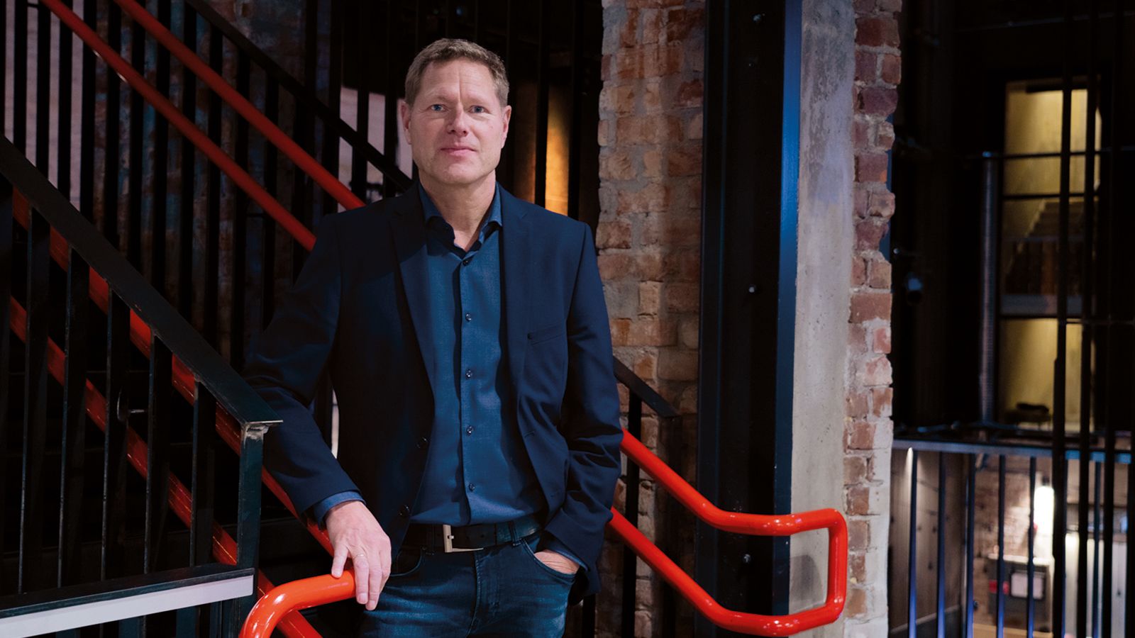 Sveinung Skule står innendørs ved et gelender i et murbygg og ser mot kamera. Store deler av bildet er dekket av en lys flate med utydelig tekst som ligger over motivet.