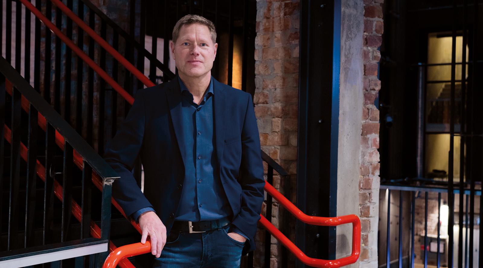 Sveinung Skule står innendørs ved et gelender i et murbygg og ser mot kamera. Store deler av bildet er dekket av en lys flate med utydelig tekst som ligger over motivet.