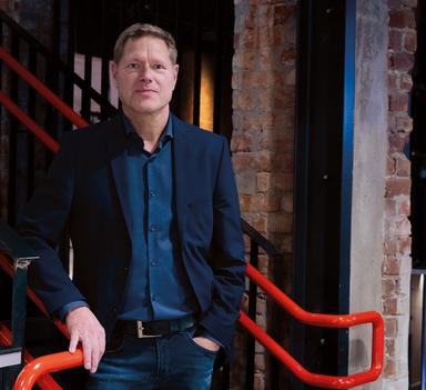 Sveinung Skule står innendørs ved et gelender i et murbygg og ser mot kamera. Store deler av bildet er dekket av en lys flate med utydelig tekst som ligger over motivet.