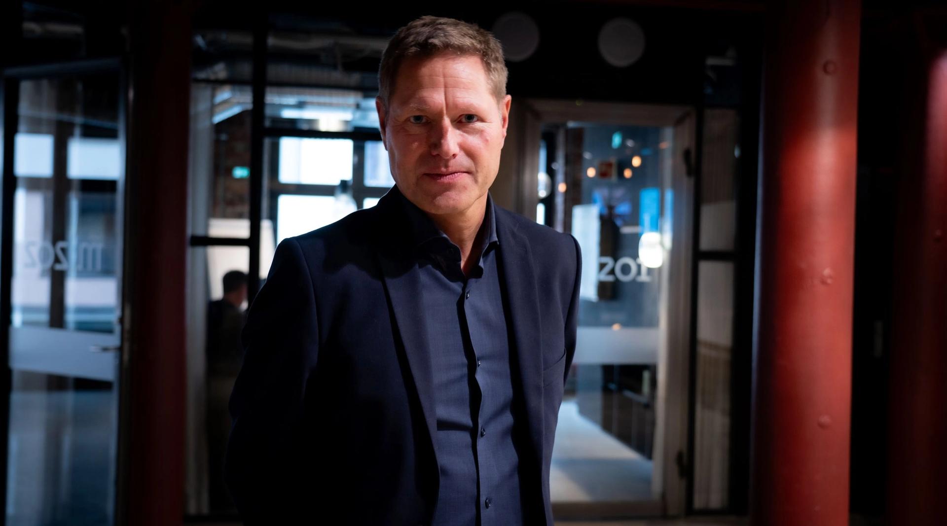 Mann i mørk dress står i et inngangsparti, bak ham er utgangsdørene i glass.
