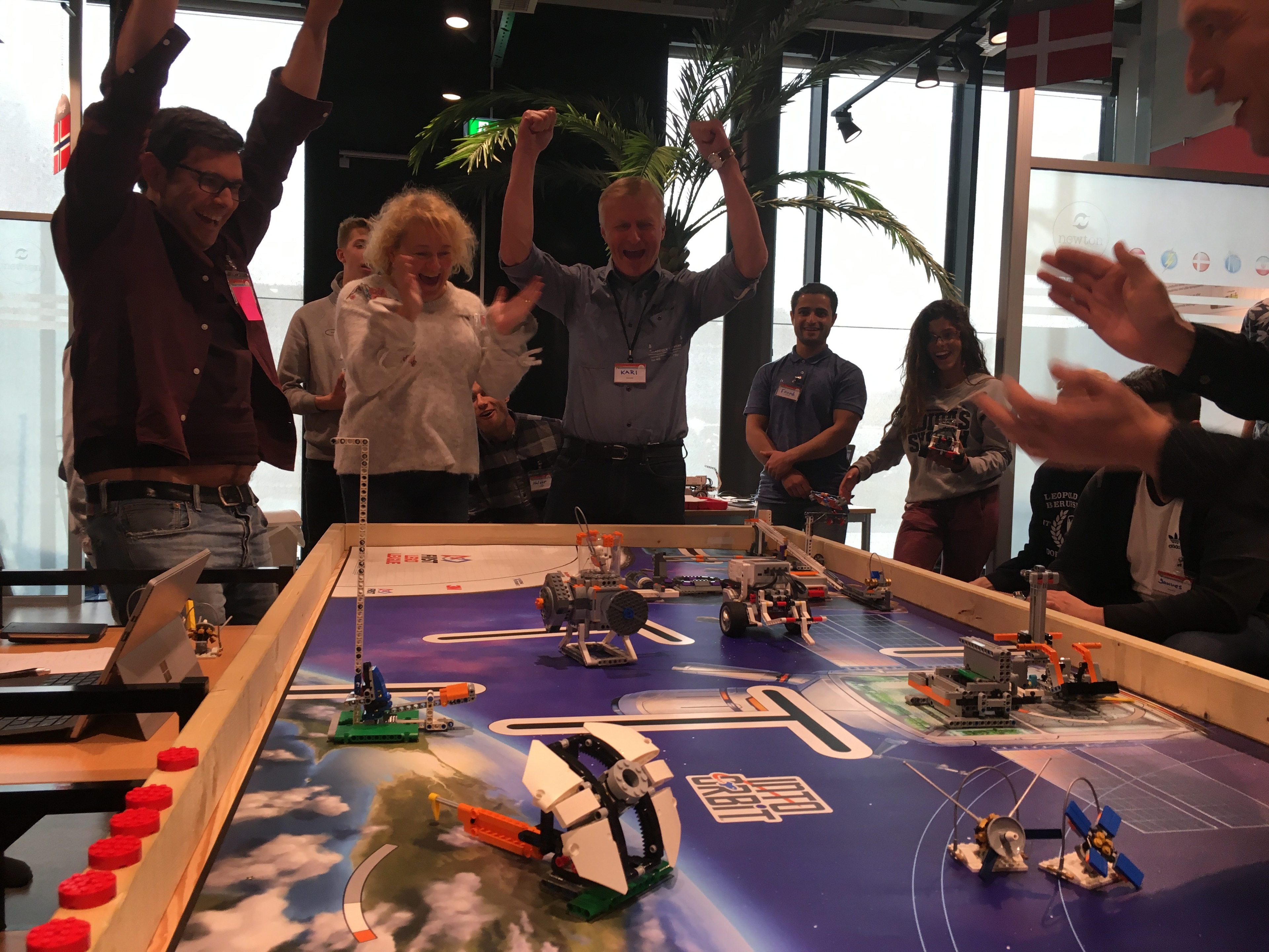 Flere lærere jubler over egne prestasjoner i en programmeringskonkurranse mens de ser på flere små roboter som kjører på et bord.