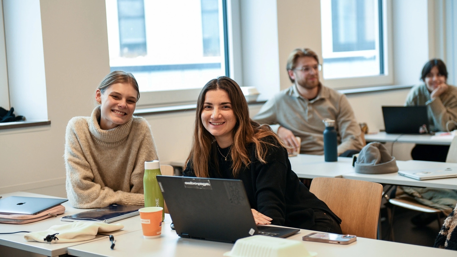 To kvinnelige studenter ser inn i kameraet i et klasserom.