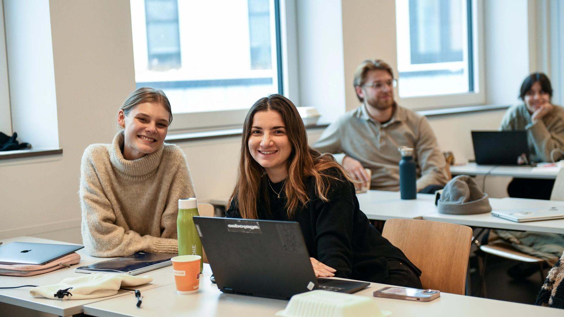 To kvinnelige studenter ser inn i kameraet i et klasserom.