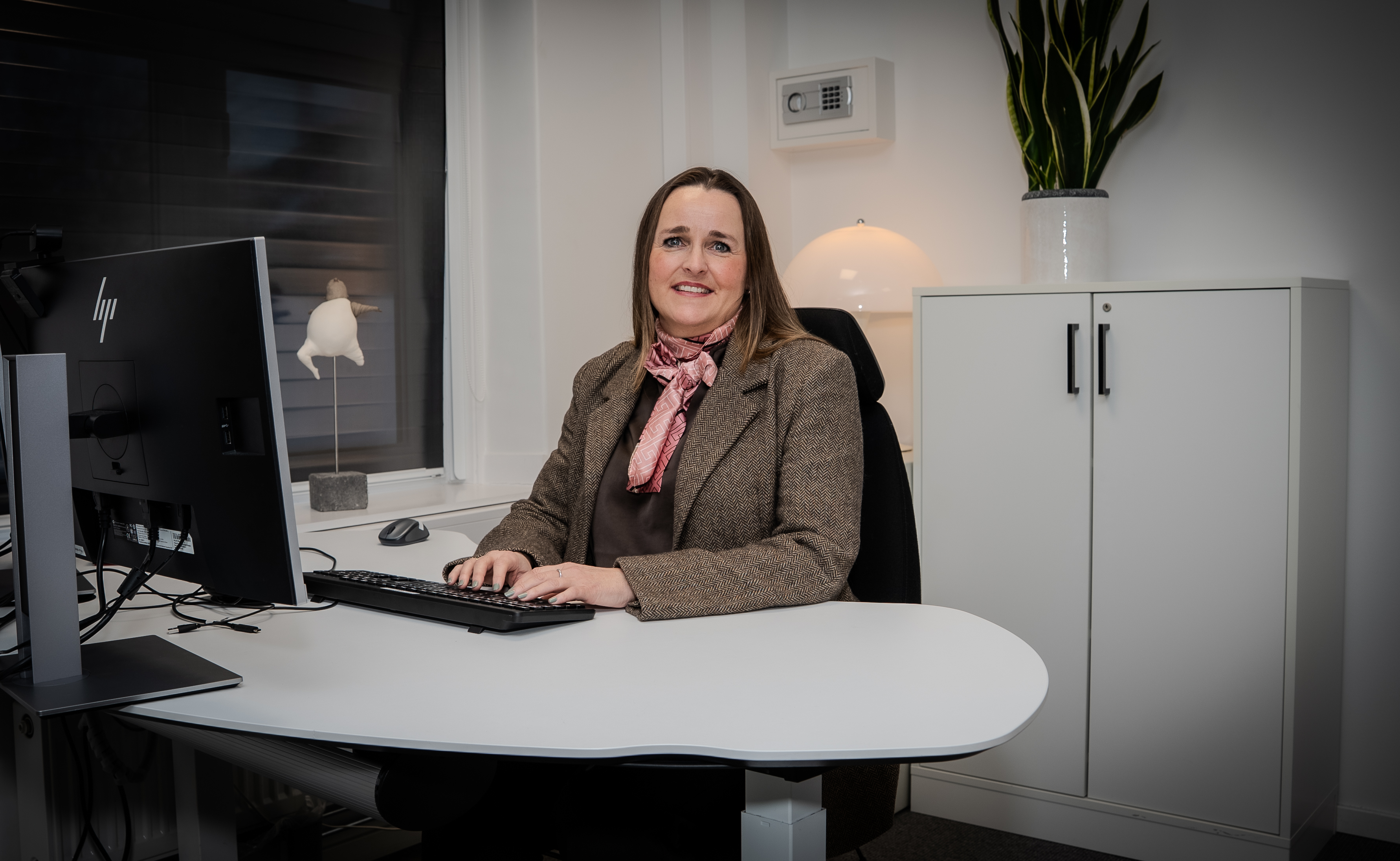 Kvinne sitter ved skrivebord på kontor og ser mot kamera. Hun jobber ved en datamaskin og har på blazer og skjerf. I bakgrunnen står et skap, en bordlampe og en grønn plante i et lyst og ryddig kontormiljø.