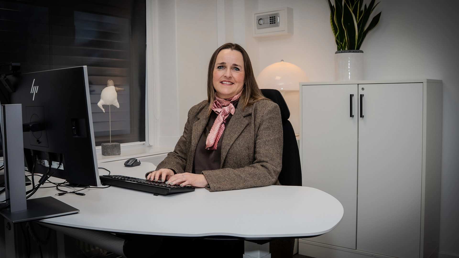 Kvinne sitter ved skrivebord på kontor og ser mot kamera. Hun jobber ved en datamaskin og har på blazer og skjerf. I bakgrunnen står et skap, en bordlampe og en grønn plante i et lyst og ryddig kontormiljø.