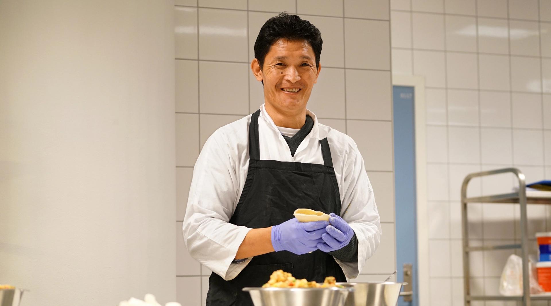 Mann i kokkeklær smiler til kamera mens han lager mat