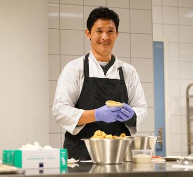 Mann i kokkeklær smiler til kamera mens han lager mat