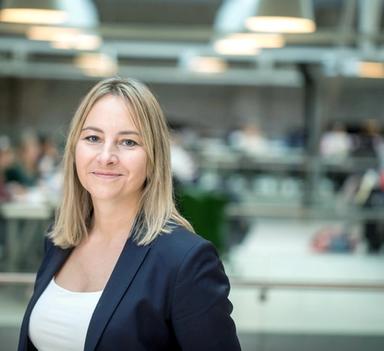 Kvinne med dressjakke ser mot kamera og smiler. Innendørs med uklar bakgrunn.